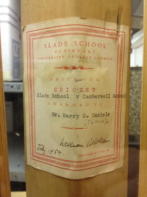 Cricket bat. A label printed with a red border and text is affixed to the bat. It begins, “Slade School of Fine Art University College London”. Below, red text, black typewritten letters, and a handwritten signature and date fill the rest of the label.