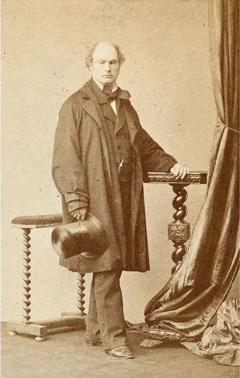 Black and white photograph. In warm tones of brown and parchment white, a pale-skinned, balding man poses in a studio. He is clean shaven and wears a baggy coat over a full suit. He holds a tall, shiny top hat in one hand, and the other rests on a tall stand with a table-top. 