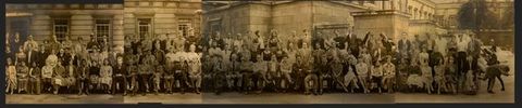 Black and white photograph. Around 125 men in and women stand or sit in three long lines in this class photograph. Many have pale skin but some men and women, the women wearing saris, have darker skin. Some smile, some look serious, and most mug for the camera. One man in a wheelchair holds a rope attached to another man, who pretends to flee to our right.