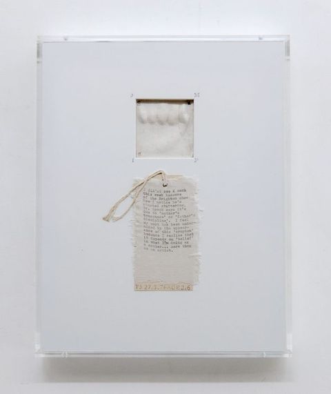 Mixed media. A square window is cut near the top centre of a rectangular white card to reveal what might be a plaster cast of a baby’s hand. Words are typewritten on a swatch of cotton fabric below. The fabric is affixed to a tag with twine strung through a hole at the top.